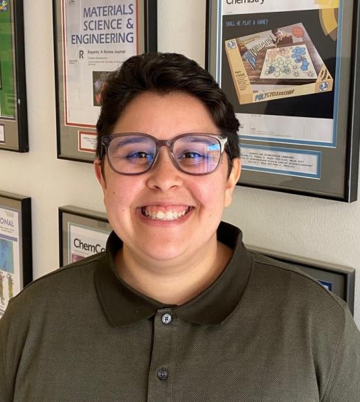 Sarah is wearing an olive green polo shirt, glasses, and a short haircut. Sarah is standing in front of a wall of old chemistry journal articles.