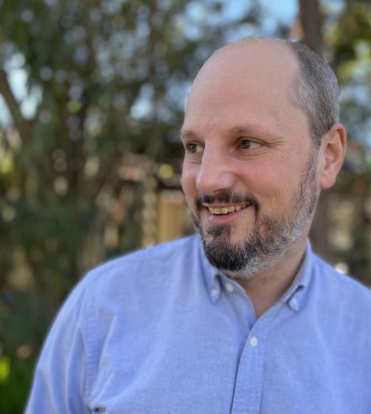 Photograph of smiling bearded man in blue shirt against blurred green foliage